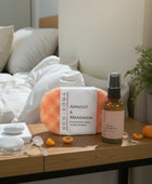 Homaroma Apricot & Mandarin exfoliating soap sponge and pillow mist set on a wooden nightstand with halved apricots.