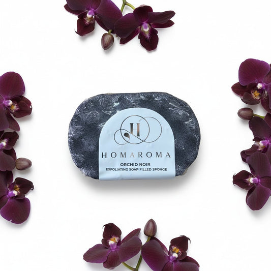 Homaroma Orchid Noir exfoliating soap-filled sponge with a dark grey textured surface, surrounded by dark purple orchids.